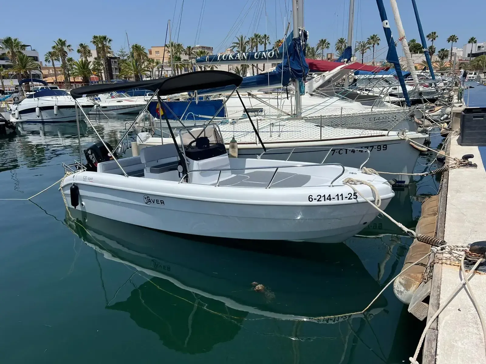 Lancha blanca atracada en un puerto deportivo, con toldo negro. Palmeras y veleros al fondo.