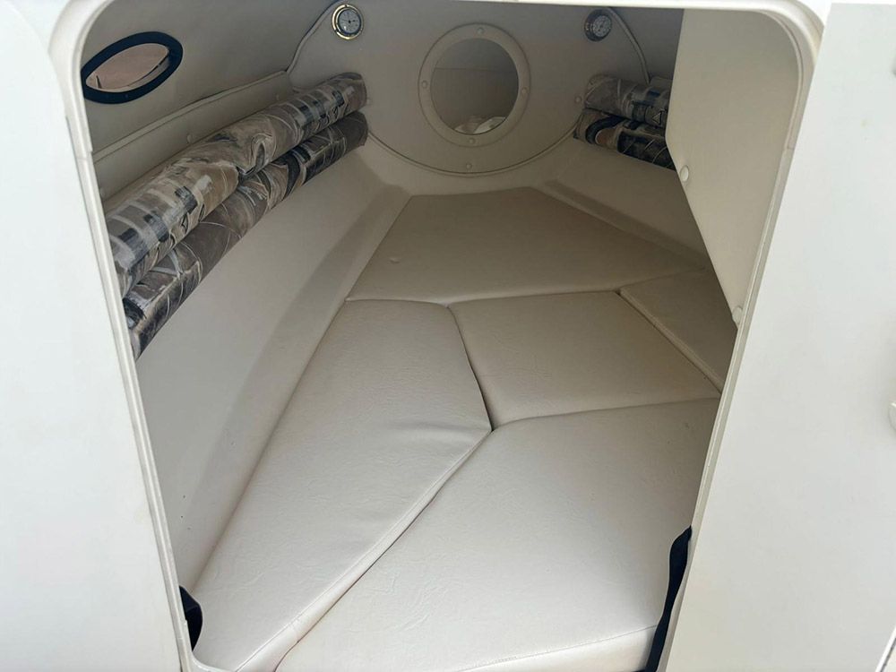 Litera blanca acolchada dentro de una pequeña cabina de barco con ojo de buey redondo y estante de almacenamiento a la izquierda.