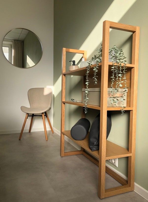 Holzregal mit Pflanzen und schwarzen Gegenständen neben beigem Stuhl und rundem Spiegel an grüner Wand