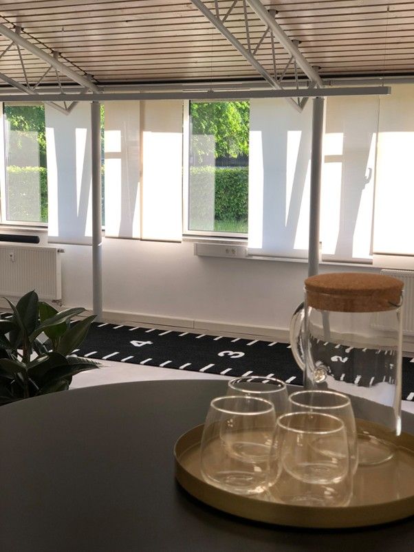 Glasflasche mit Korkdeckel und drei Gläsern auf rundem Tablett in hellem Raum mit großen Fenstern und weißen Rollos