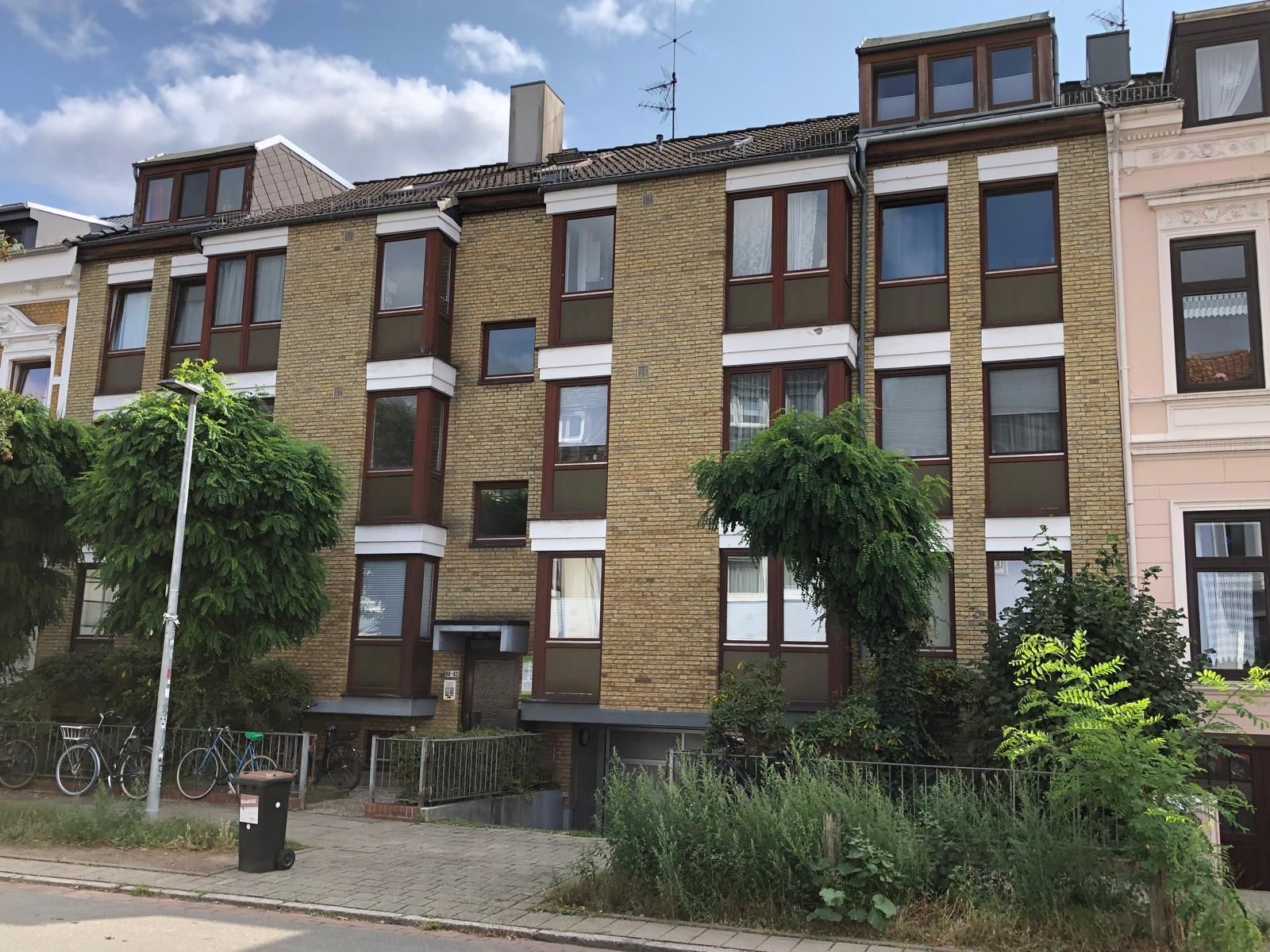 Dreistöckiges Backsteingebäude mit mehreren Fenstern, darunter Erkerfenster, und einem Flachdach; davor Bäume und Sträucher.