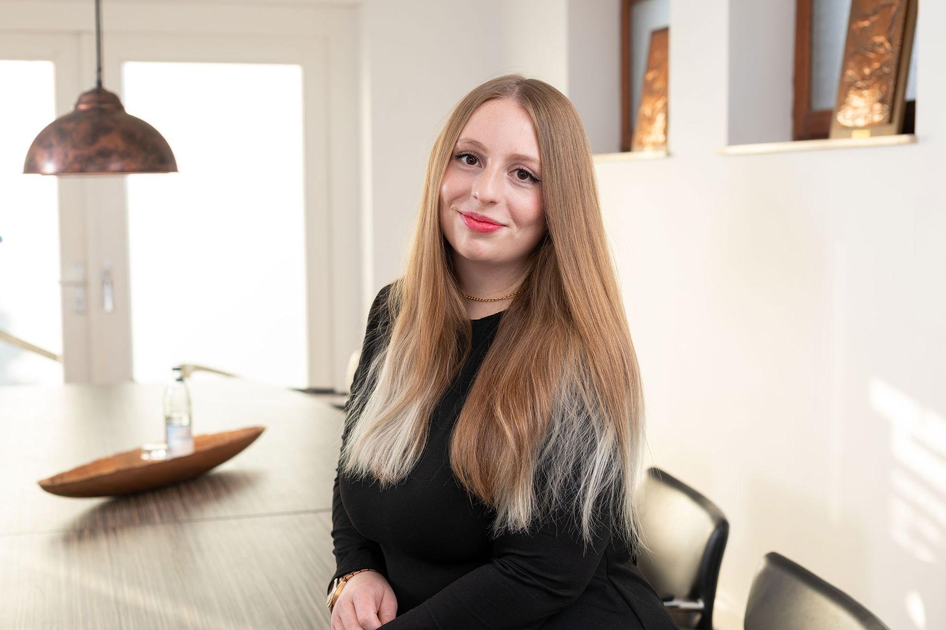 Eine Frau mit langen blonden Haaren lächelt an einem Tisch sitzend in die Kamera. Raumszene, schwarzes Oberteil.