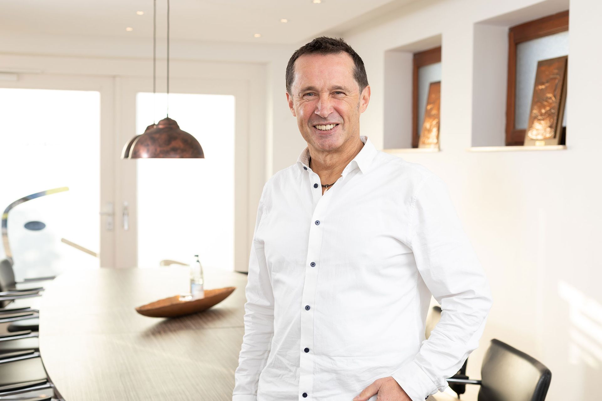 Ein Mann in weißem Hemd lächelt und steht neben einem langen Tisch in einem modernen, gut beleuchteten Raum.