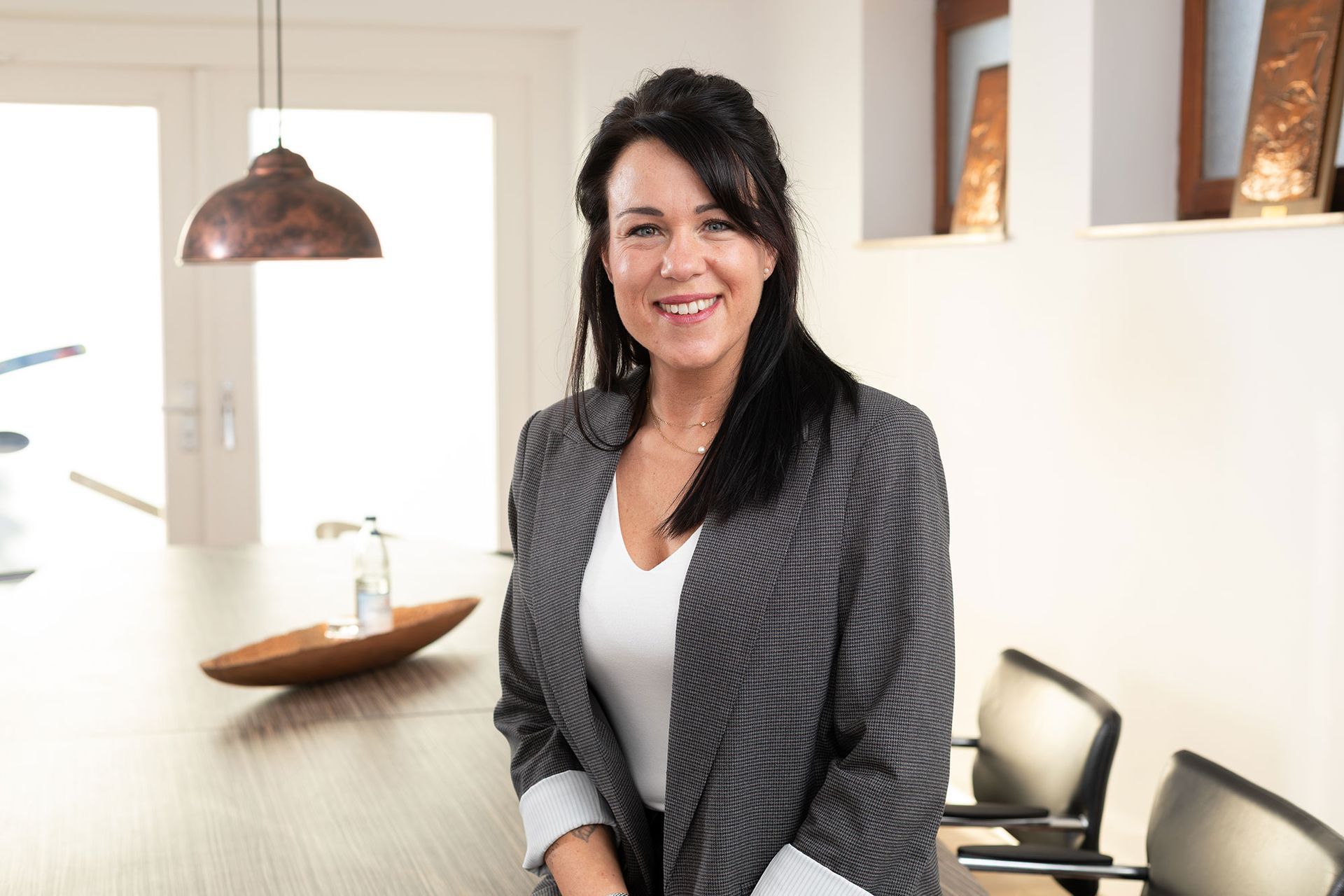 Frau im Blazer lächelt, lehnt an einer Theke; im Hintergrund ist eine moderne Büroeinrichtung zu sehen.