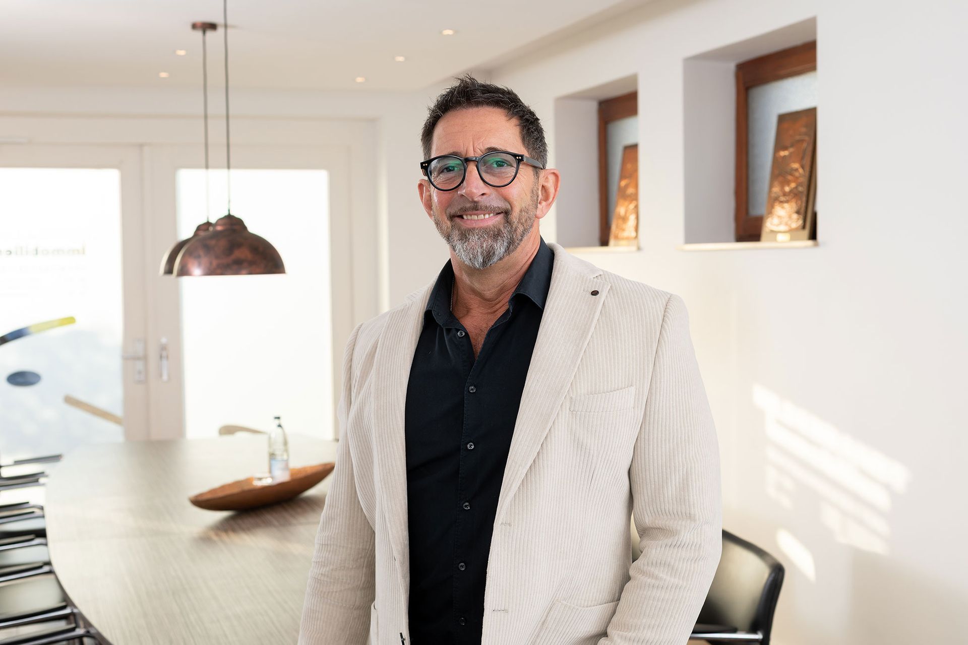 Ein Mann in Blazer und Brille lächelt und steht in einem modernen Interieur neben einem langen Tisch mit einer Hängelampe.