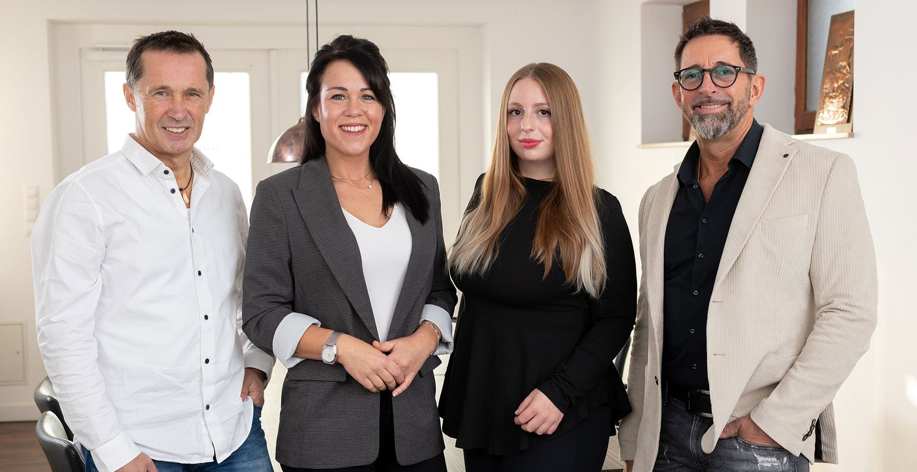 Vier Personen, zwei Männer und zwei Frauen, stehen drinnen und lächeln; neutrale Gesichtsausdrücke.