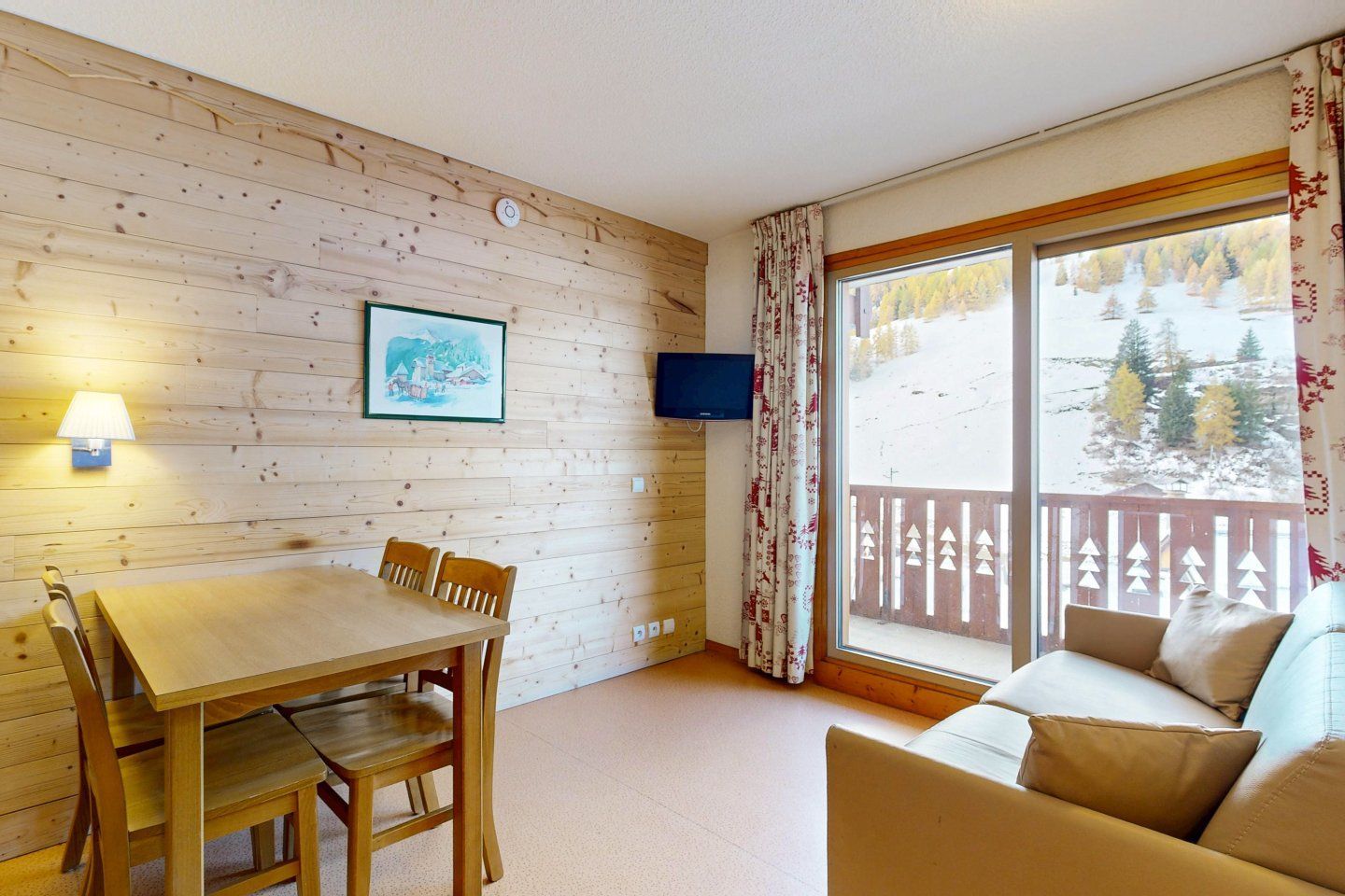 Cozy living space in a ski resort. Wood-paneled walls, dining table, sofa, balcony with a snowy view.