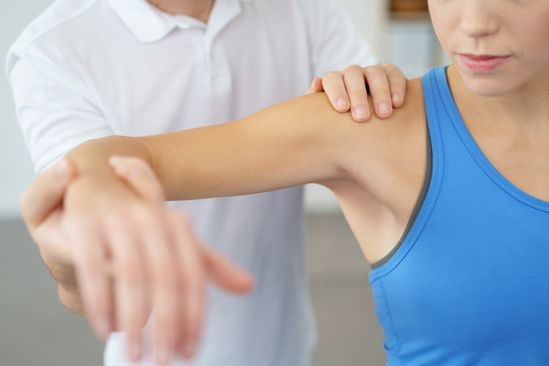 A una mujer que lleva una camiseta azul sin mangas un médico le examina el hombro.