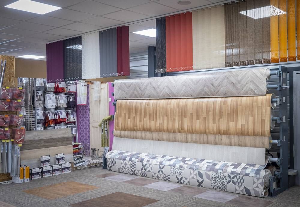 Présentation de tissus et de tapis dans une allée de magasin, avec des tapis beige, gris et à motifs enroulés sur des étagères.