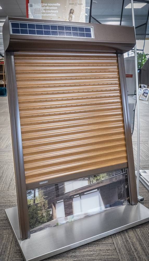 Porte de volet roulant motorisé couleur beige, avec un petit panneau solaire au-dessus, présentée à l’intérieur d’un cadre métallique.