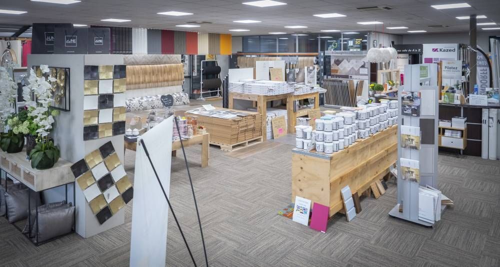 Allée d’un magasin de bricolage avec des étagères garnies de boîtes et de carrelage, ainsi que des tables d’exposition dans un espace de présentation.