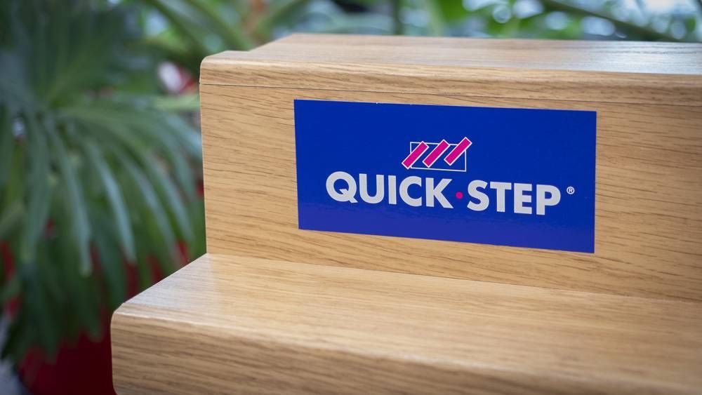 Logo QUICK-STEP bleu sur une marche en bois, avec une plante floue en arrière-plan