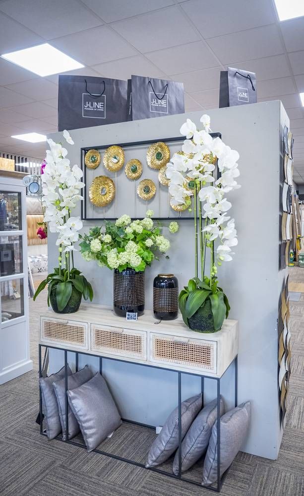 Mise en scène décorative avec des orchidées blanches, des décorations murales dorées, des bougies et des coussins gris sur une étagère de salle d’exposition