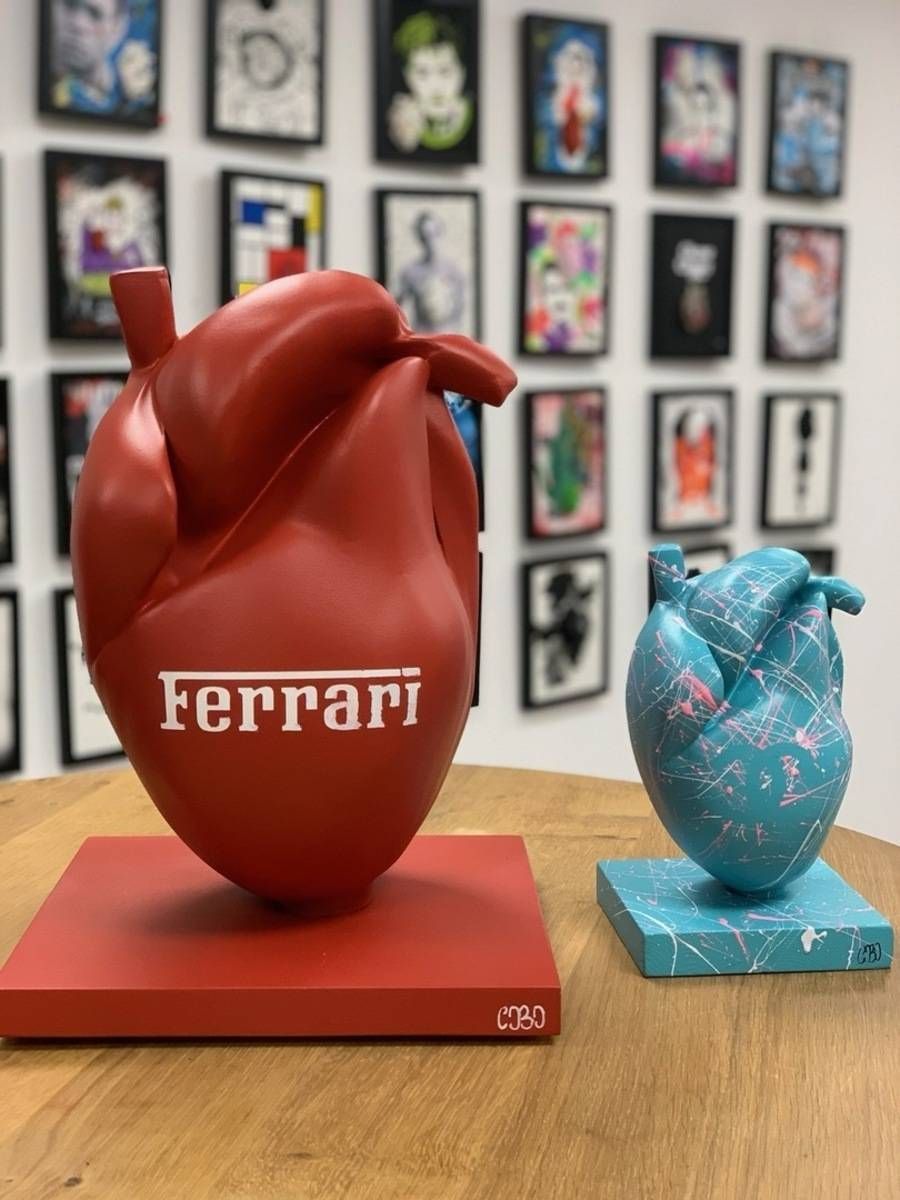 Deux sculptures en forme de cœur sur une table, l’une rouge Ferrari et l’autre bleue, avec des œuvres encadrées accrochées au mur derrière elles