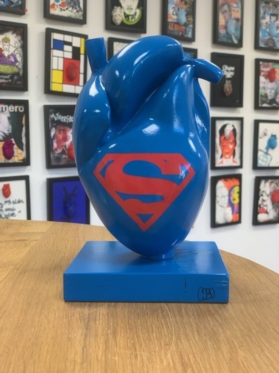 Sculpture bleue en forme de cœur, ornée d’un logo Superman rouge et posée sur un socle bleu, exposée sur une table en bois.