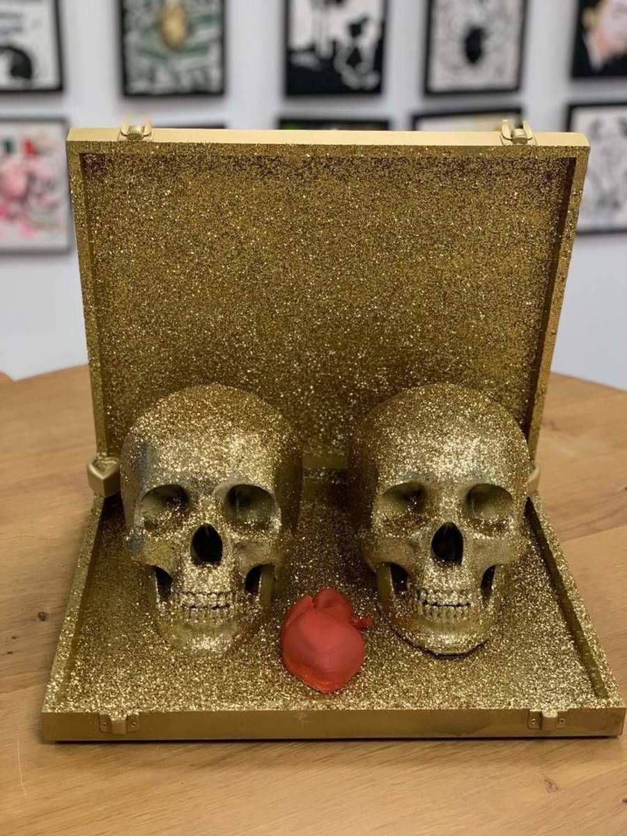 Boîte ouverte recouverte de paillettes dorées, contenant deux crânes et un petit objet rouge.
