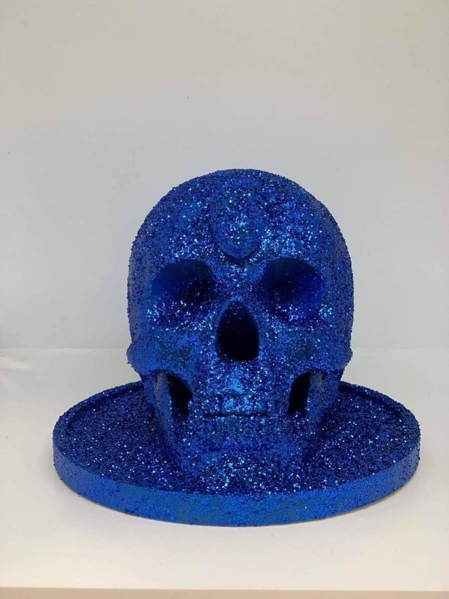 Objet en forme de crâne recouvert de paillettes bleues, posé sur un socle rond, sur fond uni