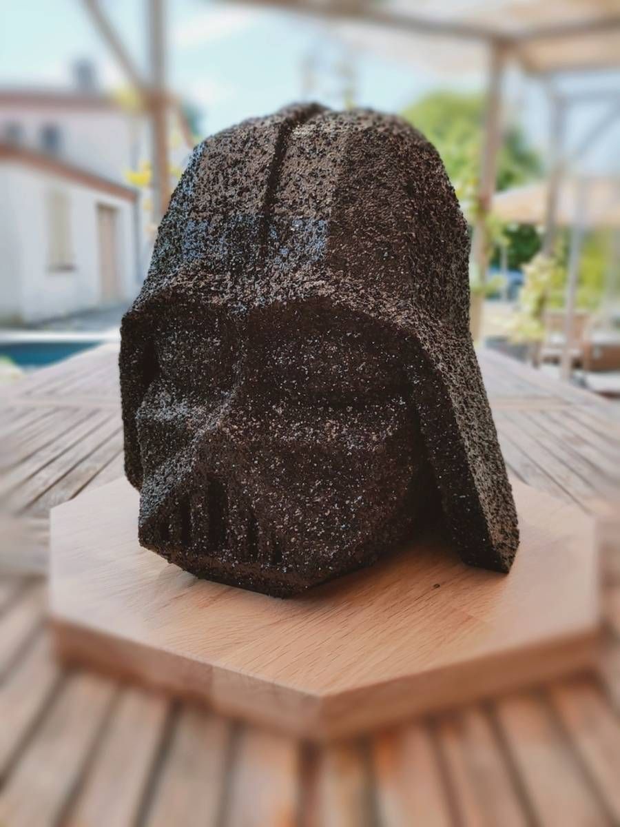 Sculpture d’un casque noir à capuche sur un socle en bois, en extérieur, avec un front de mer flou en arrière-plan
