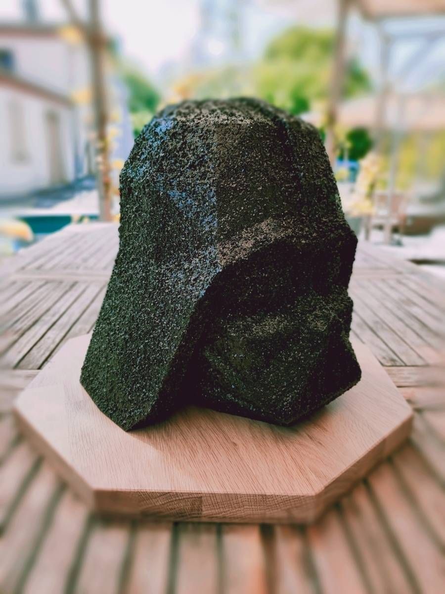 Sculpture en pierre noire à la texture rugueuse, posée sur une plateforme en bois à l’extérieur, avec une terrasse floue en arrière-plan