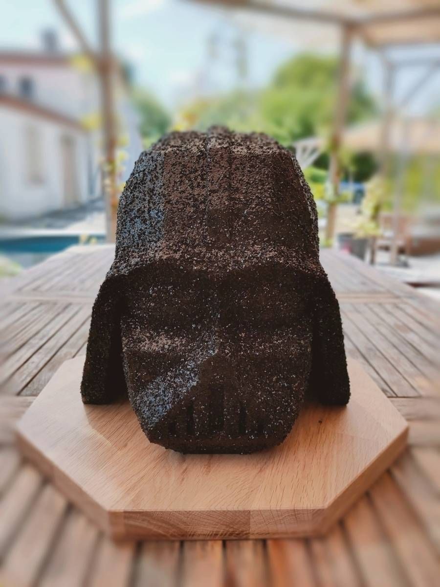 Objet sombre et texturé, sculpté, posé sur un socle hexagonal en bois à l’extérieur, avec un jardin flou en arrière-plan
