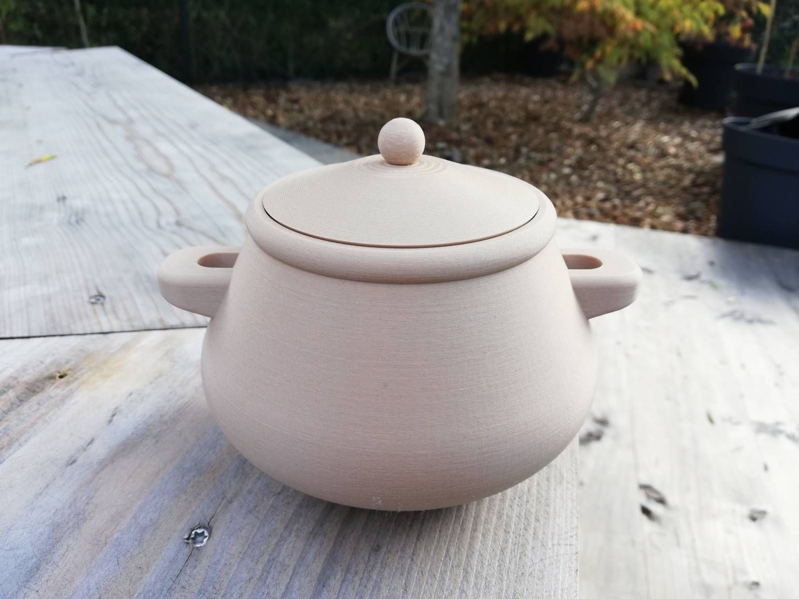 Petit pot en céramique rose pâle avec couvercle, posé sur une table en bois patinée à l’extérieur.