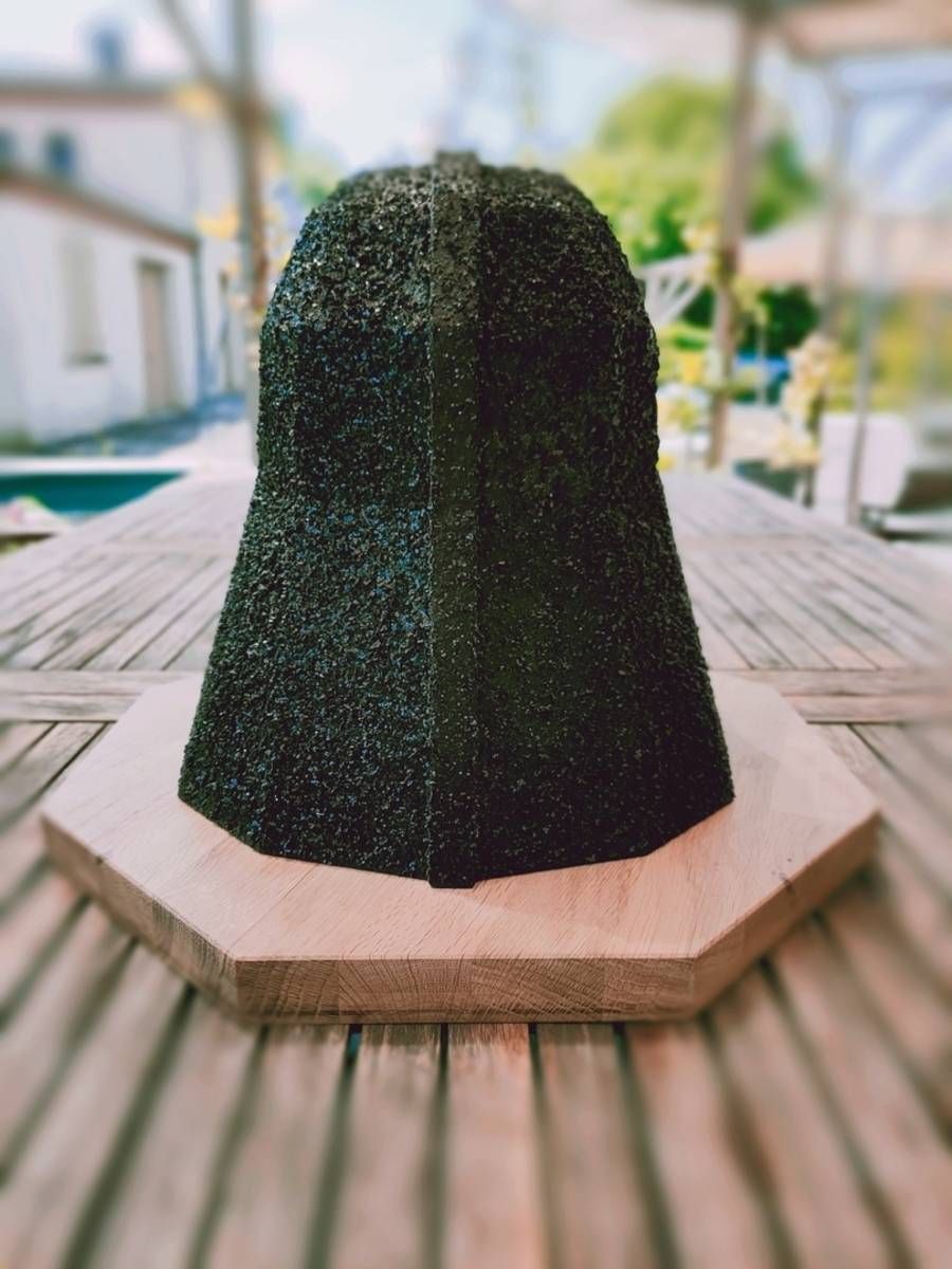 Sculpture texturée vert foncé sur un socle en pierre claire, le long d’une promenade en bois en plein air.