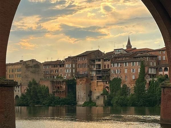 Pont de pierre au-dessus d’une rivière, avec de vieux bâtiments en briques et un clocher d’église au coucher du soleil