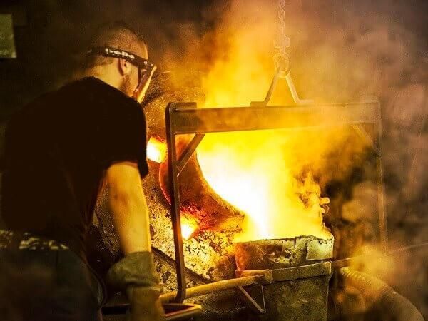 Un ouvrier s’occupant d’un four incandescent, tandis que des flammes et de la fumée s’élèvent du métal brûlant dans un atelier sombre