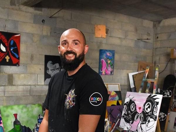 Un homme souriant en T-shirt noir se tient dans un atelier d’artiste, entouré de peintures colorées et de murs en parpaings