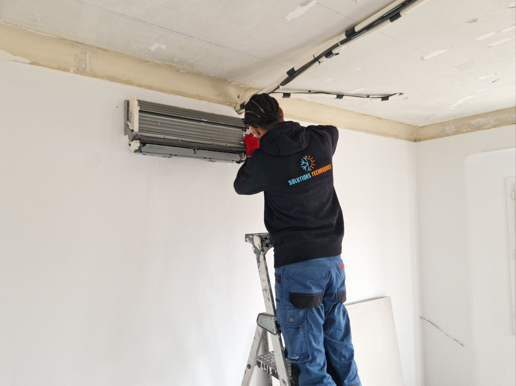 Une personne sur une échelle effectue la maintenance d'un climatiseur fixé sur un mur.