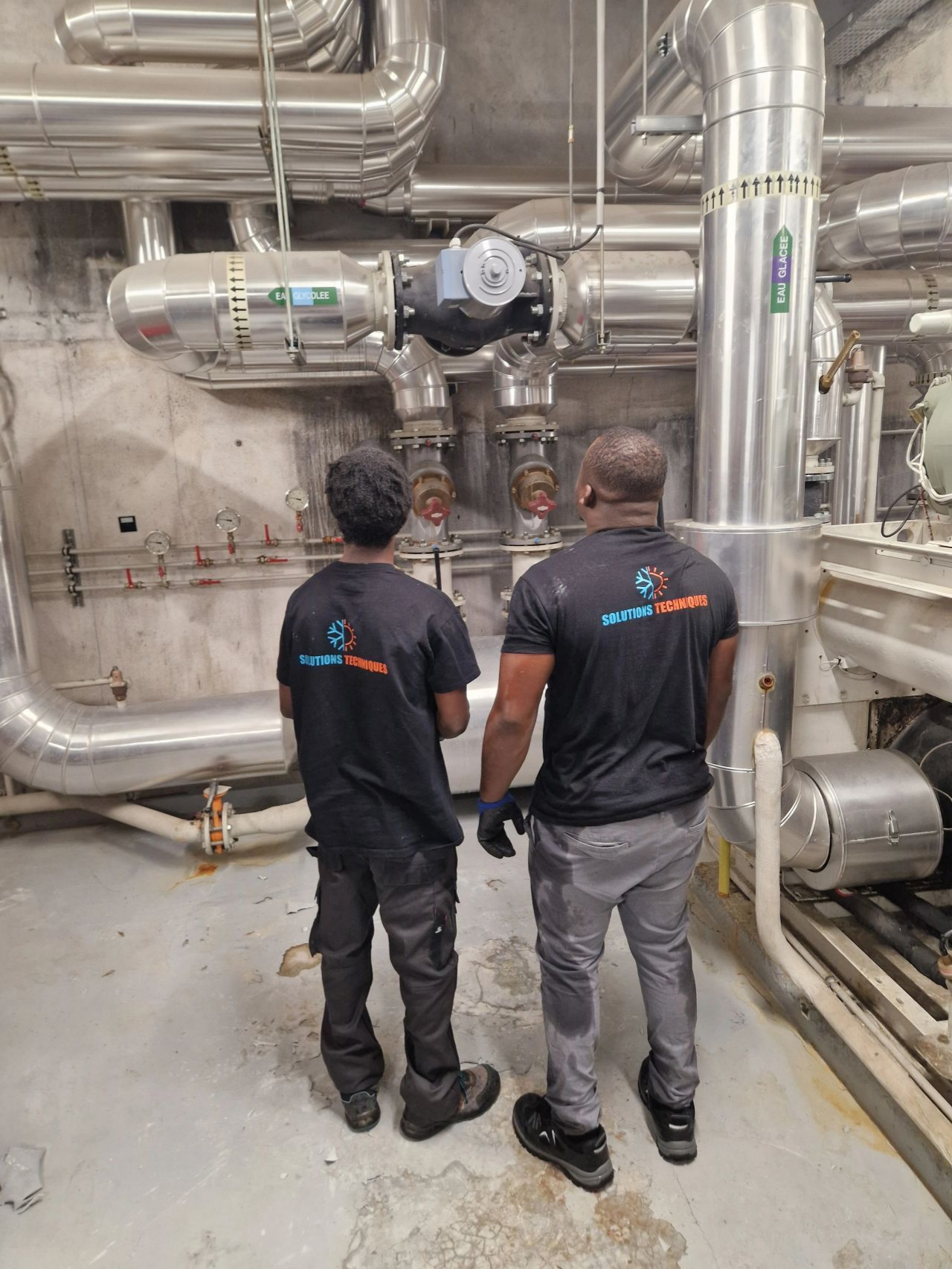 Deux personnes en tenue de travail observent des canalisations industrielles.