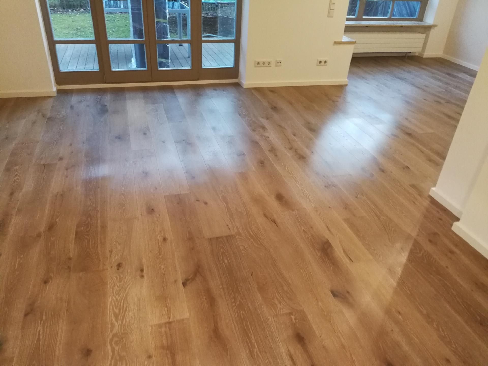 Frisch verlegter Holzfußboden in einem leeren, hellen Raum mit großen Fenstern.