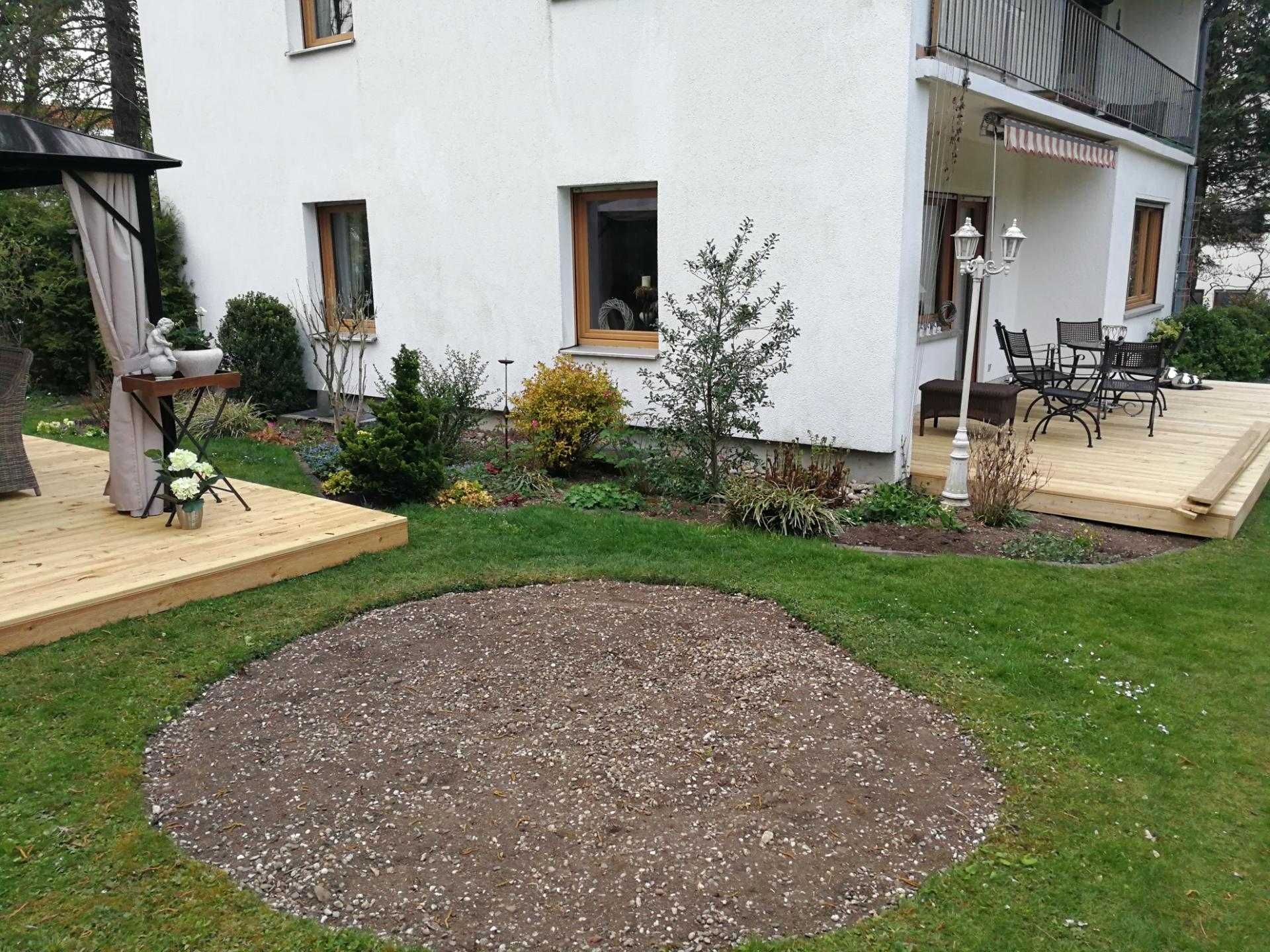 Haus mit weißer Fassade und zwei Holzterrassen, Garten mit Rasen und rundem, kahlem Erdbeet