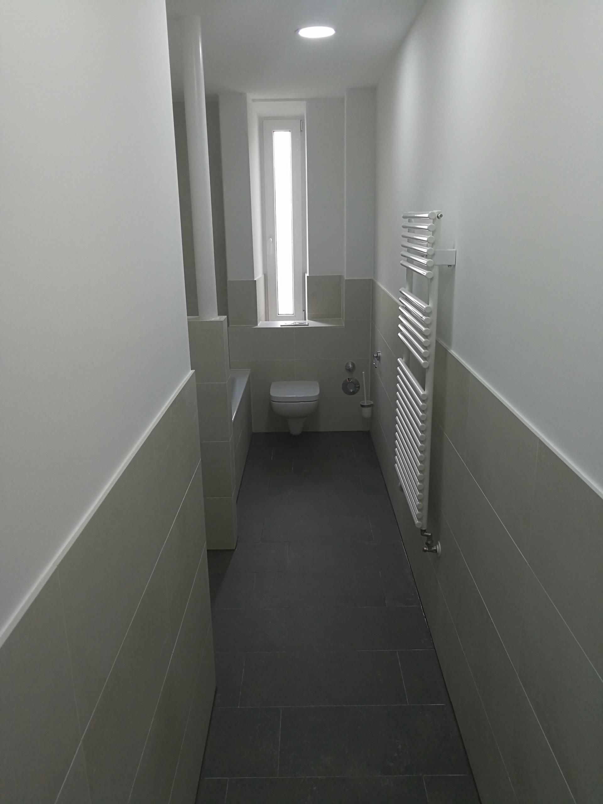 Schmaler Sanitärraum mit Toilette, grauem Boden und Handtuchheizkörper.