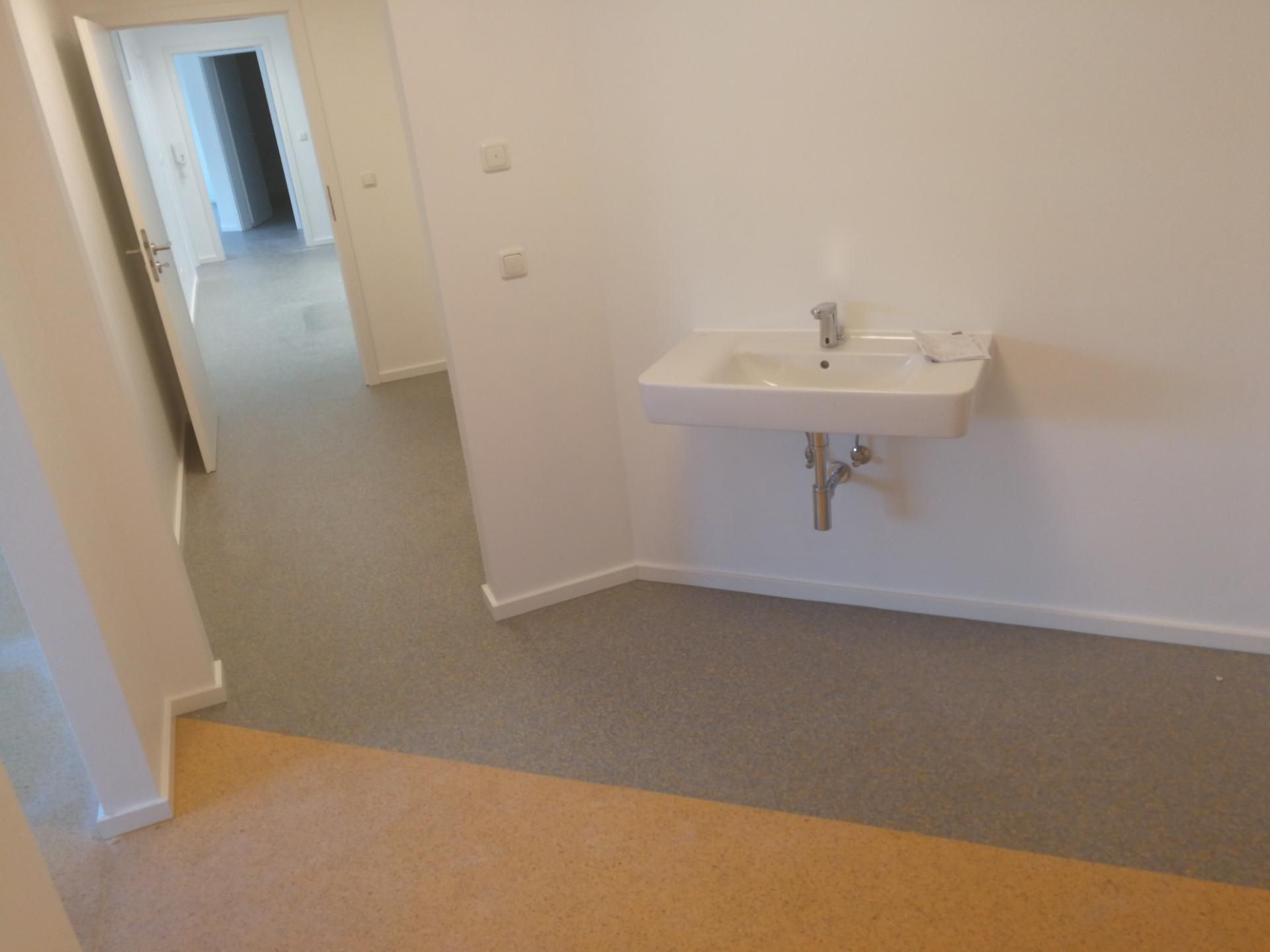 Leerer Raum mit grauem und beigem Boden, weiße Wände, Wandwaschbecken mit Wasserhahn und Seifenschale