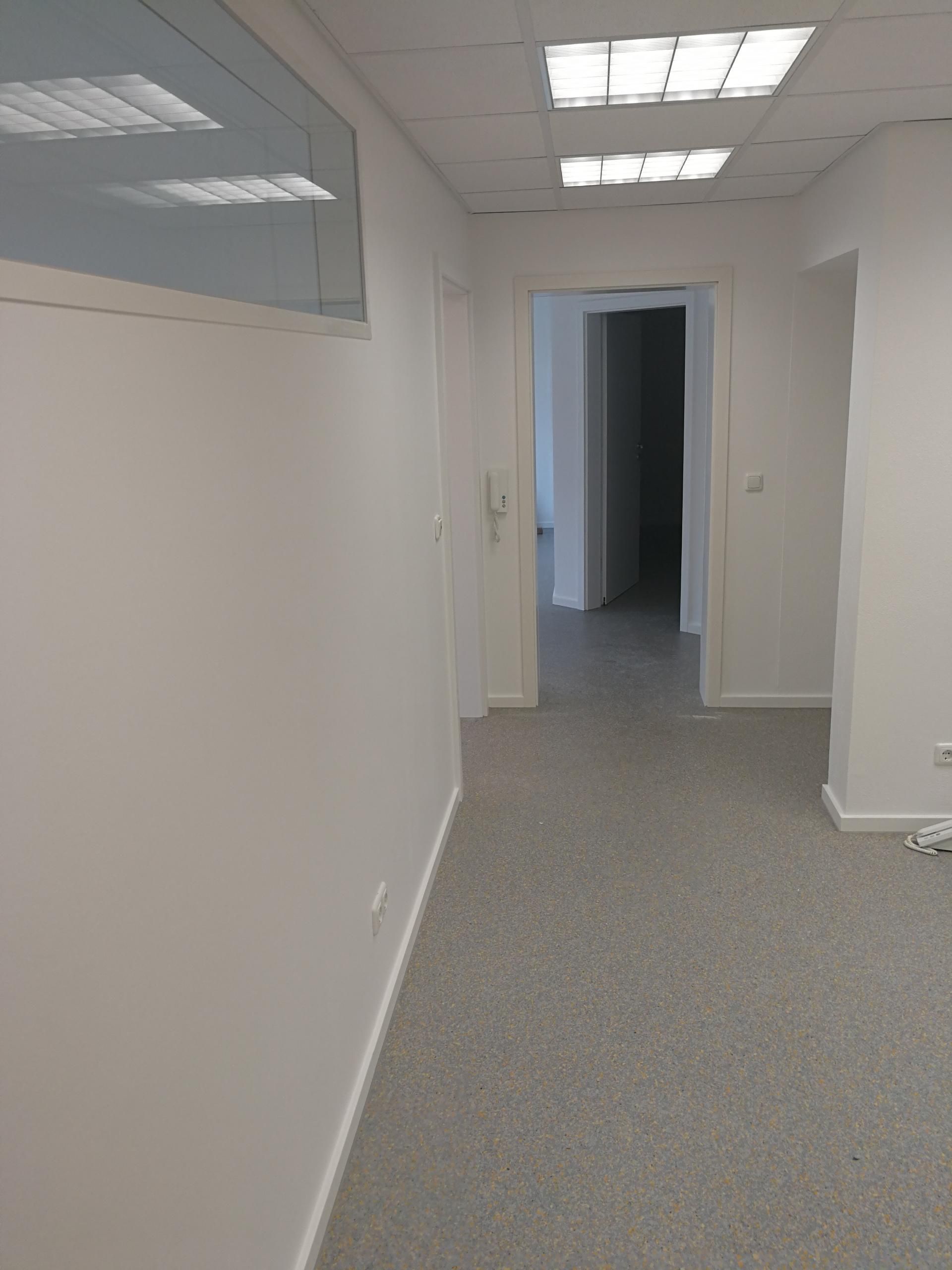 Flur mit grauem Bodenbelag, weißen Wänden und Deckenleuchten, Blick auf offene Tür zu dunklem Raum