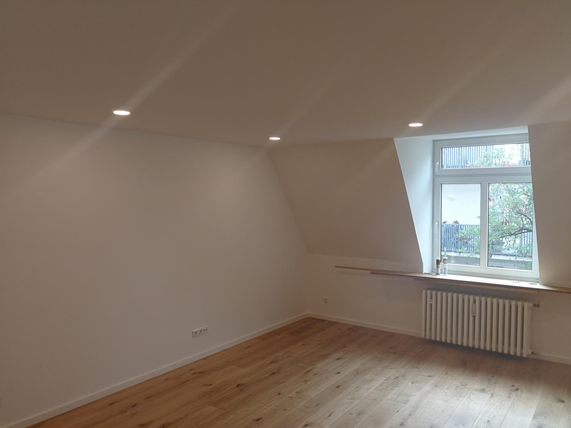 Leerer Raum mit weißen Wänden, Holzboden, drei Deckenlampen und einem Fenster mit Blick auf einen Garten