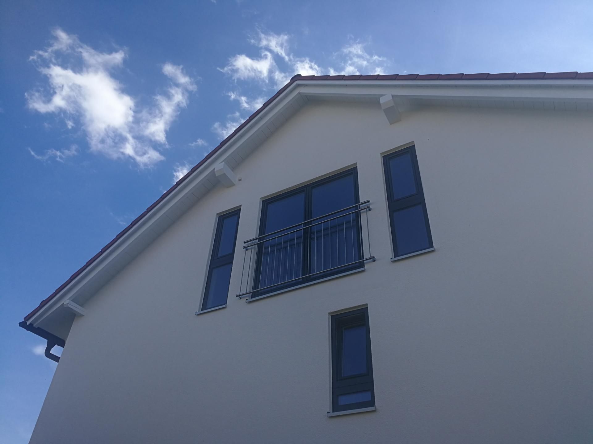 Giebel eines weißen Hauses mit drei Fenstern und kleinem Balkon unter blauem Himmel
