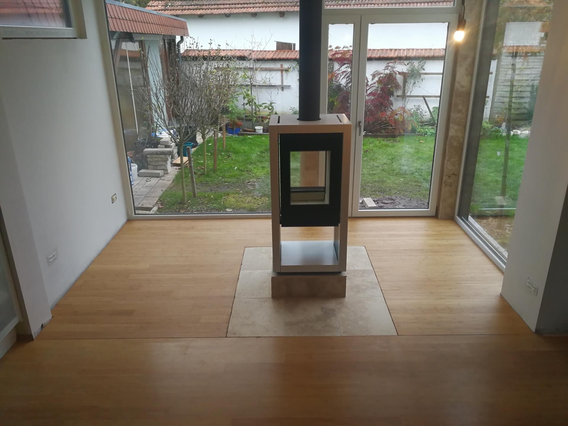 Moderner Kaminofen mit Glasfront steht auf Holzfußboden vor großen Fenstern mit Blick auf Garten