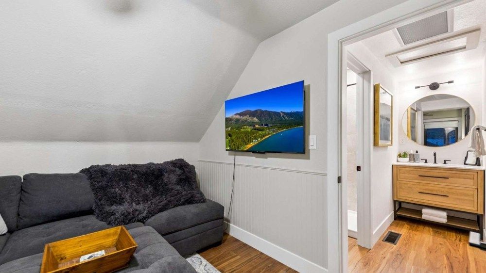 Cozy attic sitting area with gray sectional, wall-mounted TV, and hallway to a bathroom with wood vanity