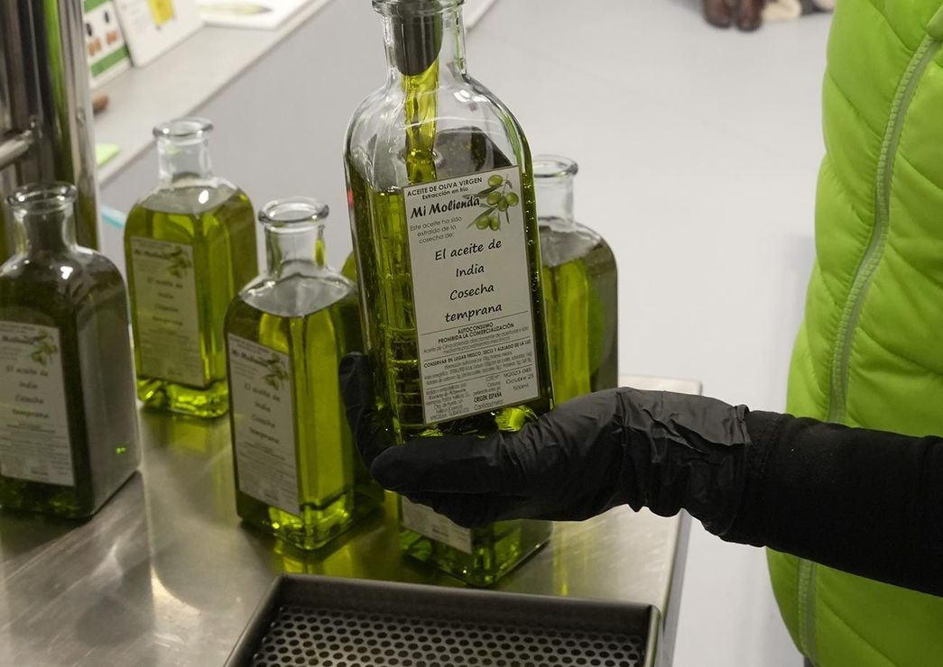 Persona con guante negro sostiene una botella de aceite de oliva, cerca de otras sobre una superficie de acero inoxidable.
