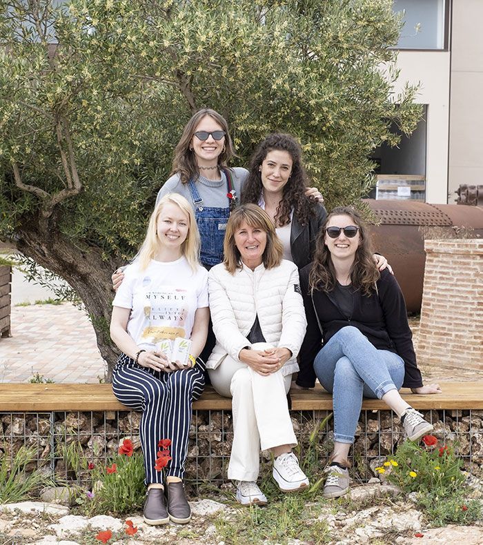 Cinco mujeres sonriendo, sentadas en un banco afuera, cerca de un árbol y una pared de ladrillos.