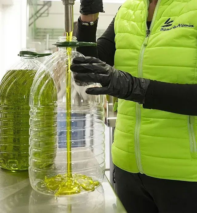 Persona con chaleco verde llenando grandes botellas de plástico con aceite de oliva; entorno industrial.