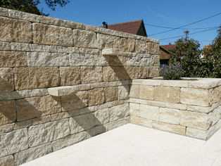 Eine Steinmauer mit einer Terrasse davor und einem Haus im Hintergrund.