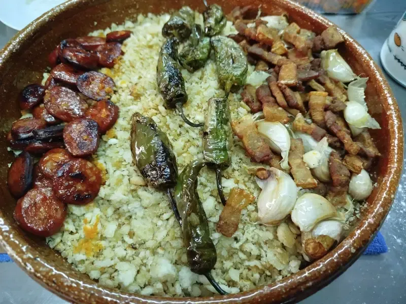Un cuenco de cerámica marrón lleno de migas, cubierto con chorizo ​​en rodajas, pimientos verdes fritos, trozos de panceta 