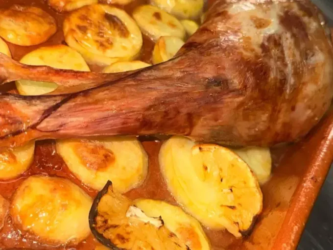 Pierna de cordero asada servida con patatas en rodajas en una salsa rojiza a base de tomate, dentro de una fuente de barro.