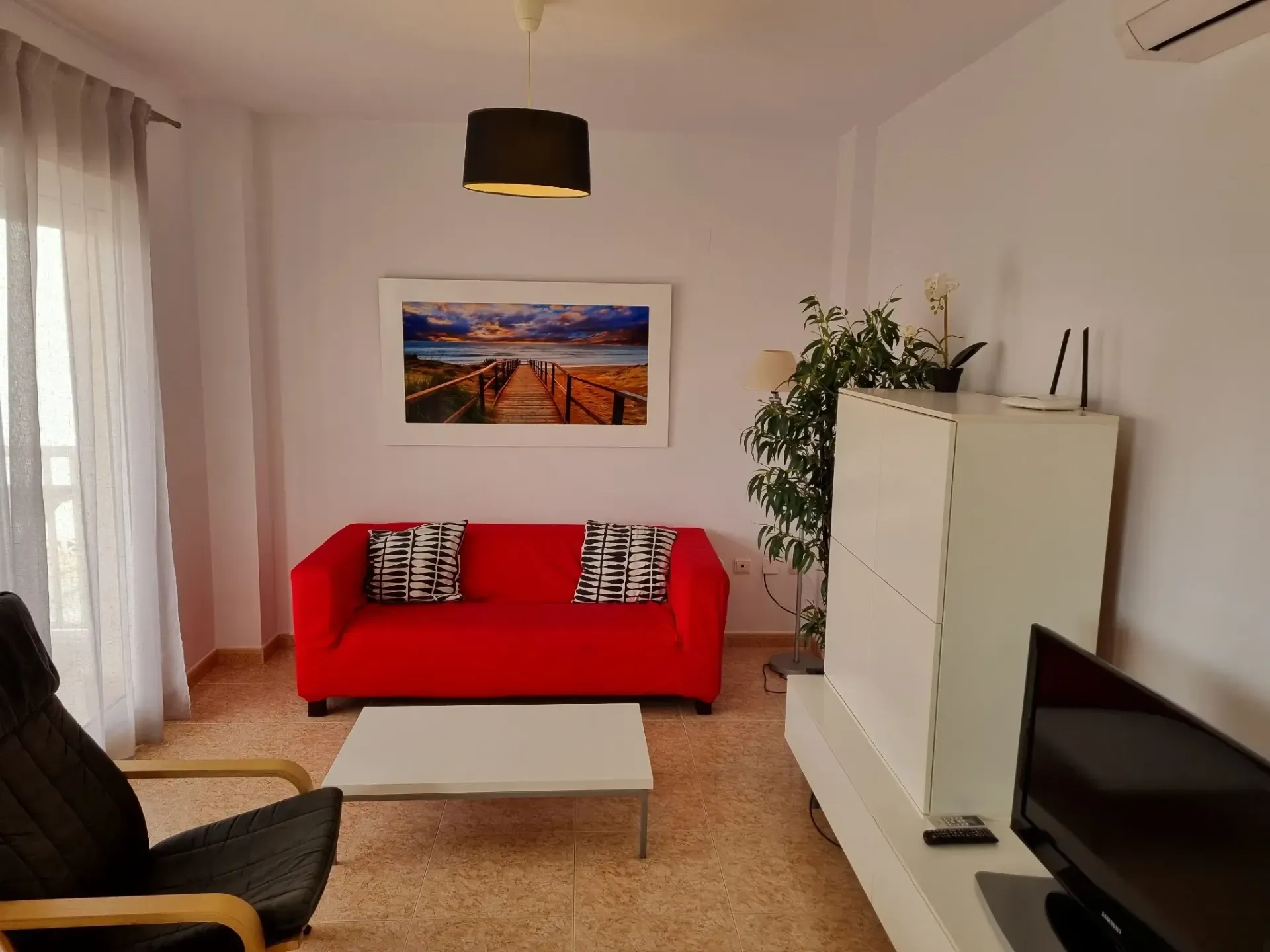 Sala de estar con sofá rojo, mueble blanco, TV y obras de arte con temática de playa.