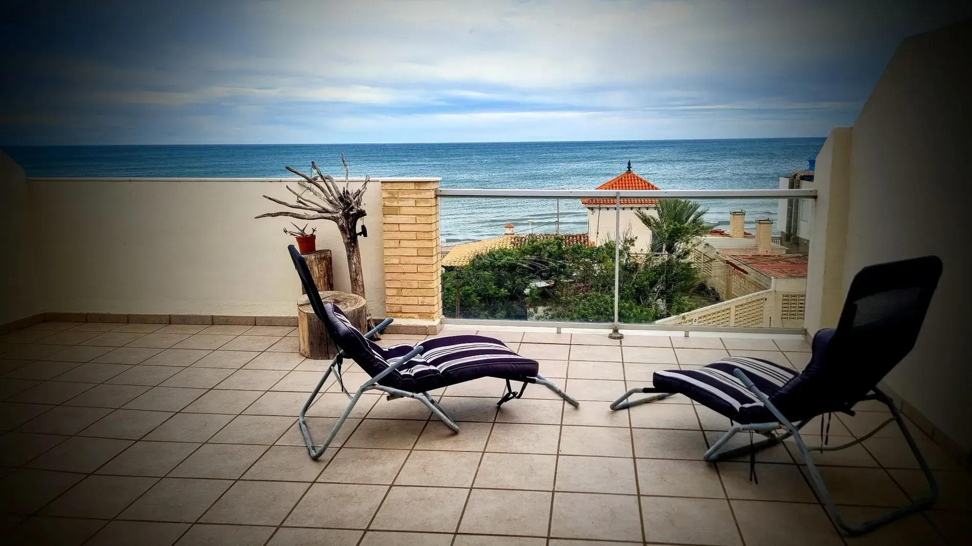 Dos tumbonas en una terraza de azulejos con vistas al océano bajo un cielo nublado.