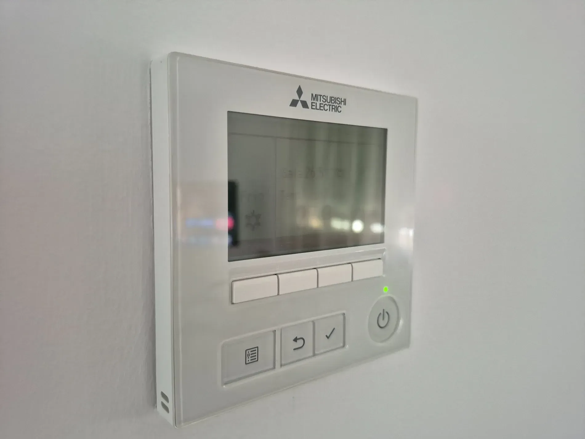 Controlador de aire acondicionado Mitsubishi montado en una pared blanca; cuenta con una pantalla y varios botones de control.