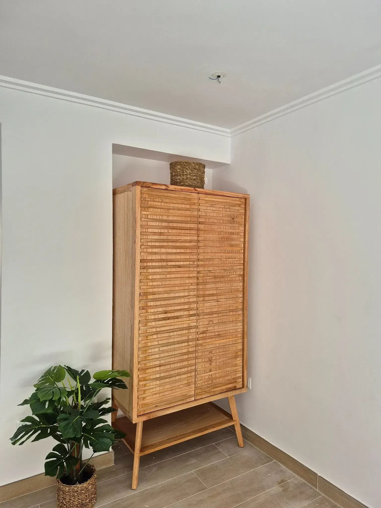 Armario de madera en un rincón de paredes blancas con una planta en una maceta tejida.